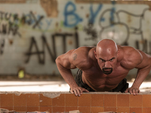 Etory, bald and muscular, leans forward with hands on a brick wall, showcasing a defined chest and intense expression. Etory's large, hairy ass is partially visible.