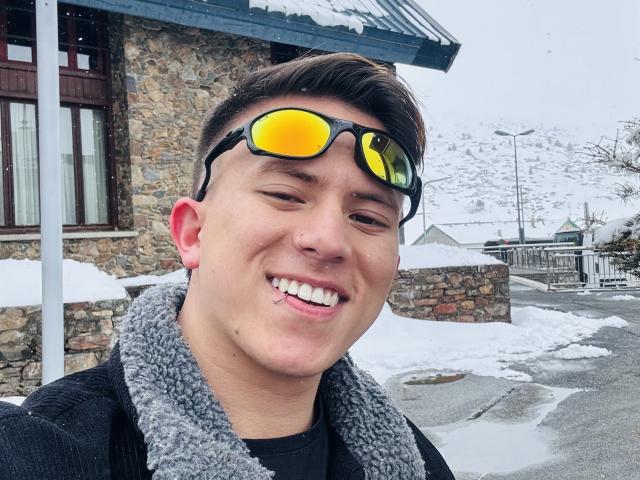 BastianC with yellow reflective sunglasses, smiling, showing a small metal stud in his bottom lip, wearing a gray fuzzy collar, standing outside in a snowy area.