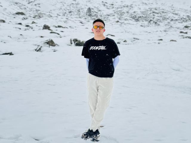 BastianC standing in snow, wearing reflective goggles, black t-shirt with white text, white pants, and black sneakers, arms behind back, short dark hair, overcast winter day.