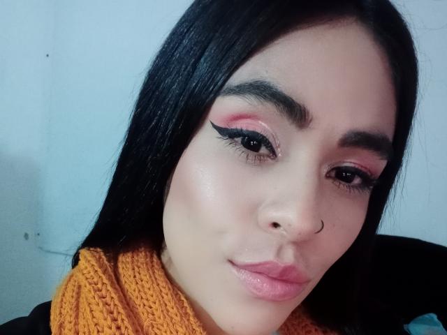 StellaBeckker has pink eyeshadow, black eyeliner, and a nose ring. She's wearing a yellow knitted scarf, with black hair and pink lips. Her skin is light.