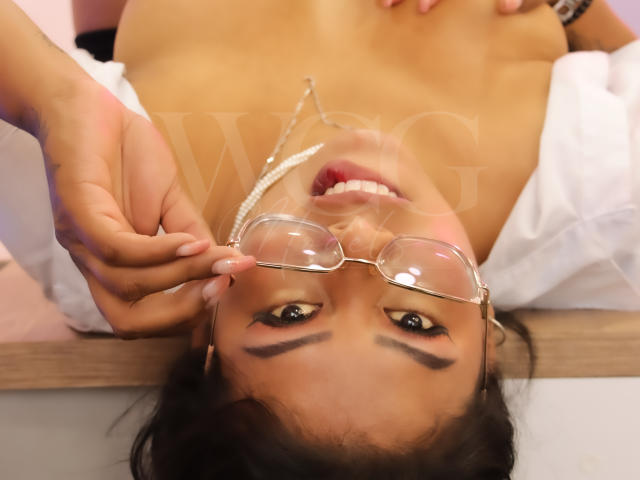 LizHerrera's tits exposed, wearing glasses, pearl necklace, lying upside down, smiling, hands adjusting glasses, white shirt partially open, brown hair, tan skin, makeup, visible teeth, relaxed.