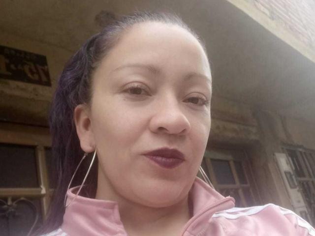  CharloteEzra with dark purple lipstick, pink track jacket, and long purple hair in a ponytail, wearing large hoop earrings, smiling slightly.