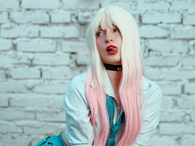 Jellyfishpink with white-to-pink gradient hair, red lipstick, black choker, white shirt, and blue dress, tongue out, looking up, against a white brick wall, in a selfie.