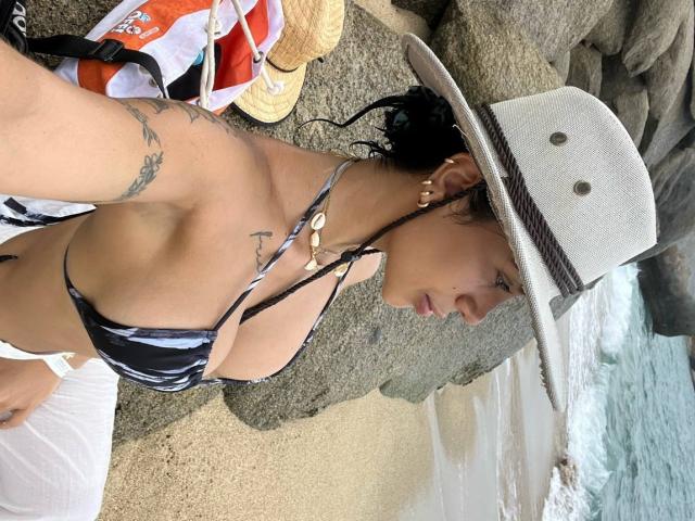 NikolRyder in a black and white bikini top, hat, and necklace, showing tattoos and small tits, laying on rocks by the ocean, with a towel and bag nearby.