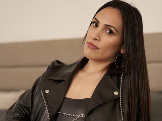 SandraHott69 with long black hair, wearing a black leather jacket and hoop earrings, has a serious expression, small tits, and a chain necklace, in a softly lit room.