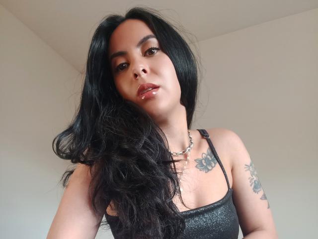 Sarah Luna, con lunghi capelli neri, labbra rosse lucide, top nero scintillante, collana a catena d'argento, tatuaggio sulla spalla destra, guarda verso il basso la fotocamera, mostrando leggermente il seno e i glutei..