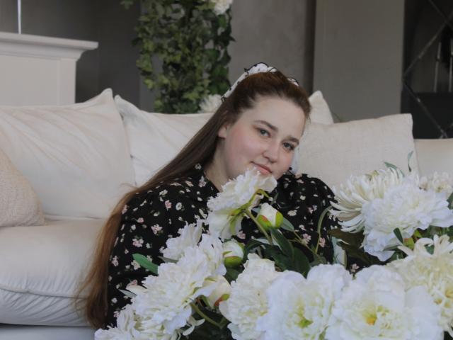 DaniellaGraceZZ with long brown hair, black floral dress, smiling while holding white peonies, sitting on white couch, white pillows, white wall in background, wearing white hair clip.