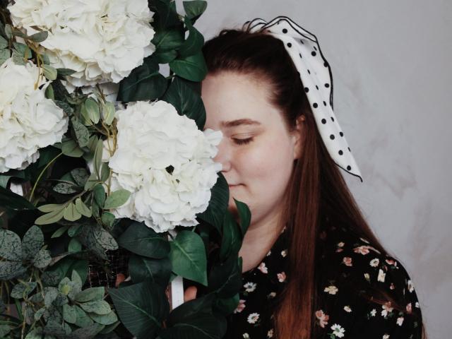 DaniellaGraceZZ with nose pressed into white hydrangeas, wearing a polka-dot headband and floral dress, eyes closed, brown hair, fair skin, and a slightly flushed face.