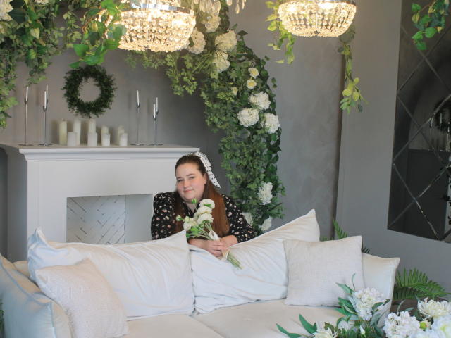 DaniellaGraceZZ, con cabello castaño, una blusa negra con lunares, sosteniendo rosas blancas, sentada en un sofá blanco, rodeada de follaje, flores blancas y candelabros de cristal..