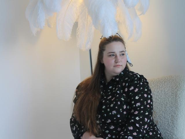 DaniellaGraceZZ has long brown hair, wearing a black floral shirt, sitting under a white feather lamp, looking slightly bored with a soft, pale skin tone.