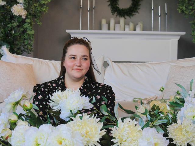 DaniellaGraceZZ has pale skin, brown hair, and is smiling with white and yellow flowers in front of her. She's wearing a black floral shirt, sitting on a beige sofa.
