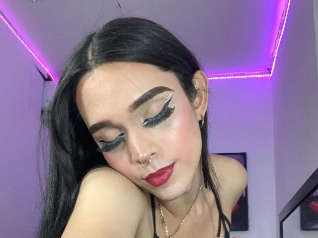 AlexaFoxxi, con capelli neri, rossetto rosso e un trucco occhi drammatico con eyeliner ad ali, un anello al naso e orecchini dorati, è in avanti, mostrando le spalle scoperte, in una stanza con illuminazione a LED viola..