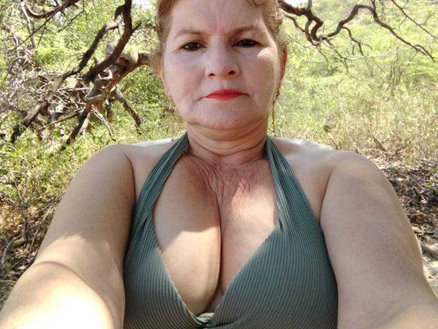 PerlaDeluxe, a middle-aged woman with red lipstick and green halter top, showing large, cleavage-filled tits, taking a selfie outdoors, with dry, twisted branches in the background.