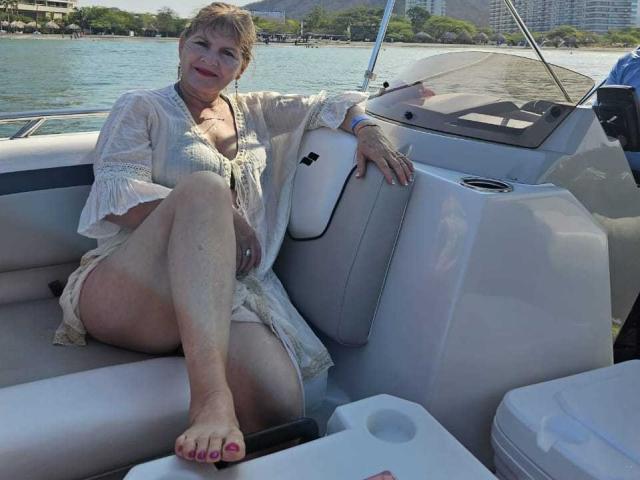 PerlaDeluxe with short blonde hair, wearing a white lace dress, sitting on a boat with her ass exposed, legs crossed, and toes painted red, smiling at the camera.