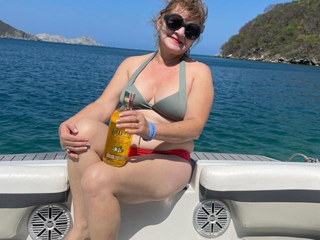 PerlaDeluxe, chubby with medium tits, wearing a gray bikini top and red bottoms, sitting on a boat, holding a yellow bottle, wearing sunglasses, with a sunny blue ocean.