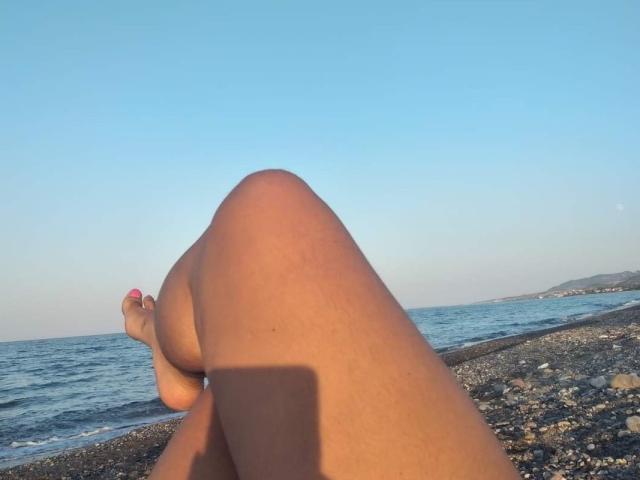 Melissac69's legs crossed, showing tan skin and pink toenails, with her feet in the foreground and a clear blue sky and ocean in the background.