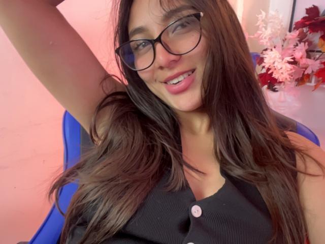 DianaFernandezz with long brown hair, glasses, black sleeveless top, smiling, arm raised, blue chair, pink wall, pink flowers in background, cute, small tits, pretty.