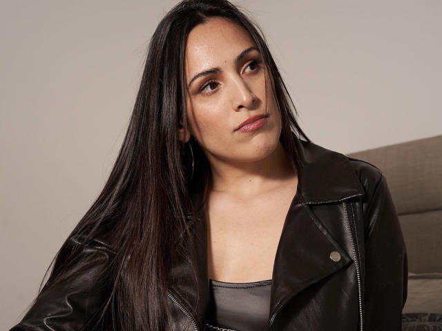 SandraHott69 with long black hair, brown eyes, wearing a black leather jacket, looking slightly to the side with a neutral expression, wearing a grey tank top.