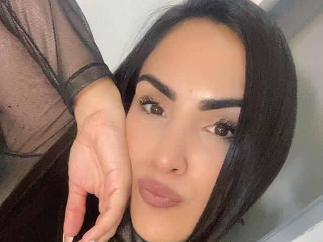 SandraHott69 with long black hair, brown eyes, and full lips, wearing sheer black top, touching her cheek with her right hand, showing subtle makeup and natural skin tone.