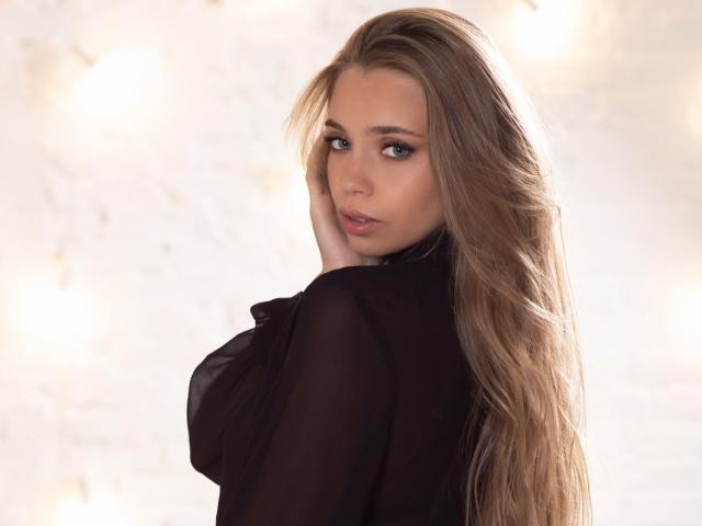 LizaSoTrue mit langen, welligen blonden Haaren, die ein durchsichtiges schwarzes Oberteil trägt, blickt über ihre Schulter mit einem leichten Lächeln und zeigt ihr dezentes Make-up und ihr zartes, hübsches Gesicht..
