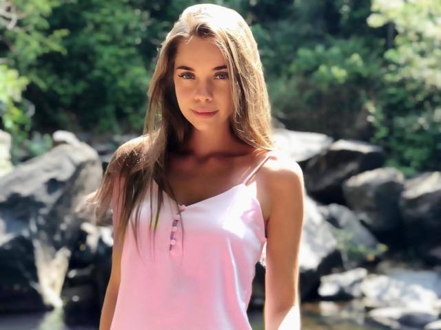 LizaSoTrue con cabello castaño largo, top rosa sin mangas, pechos pequeños, cuerpo delgado, de pie al aire libre, luz solar, fondo verde, rocas, ligera sonrisa, luz natural, atractiva, sexy..