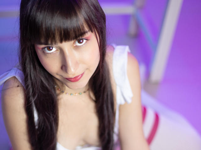 AliceLim with long dark hair, bangs, and pink eye makeup, looking up with a slight smile, wearing a white top, showing her small tits, in a purple-lit room.