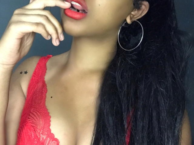 Rishna with red lips, black hair, red lace bra, and silver hoop earrings, touching her lips with her fingers, showing a small star tattoo on her right shoulder.