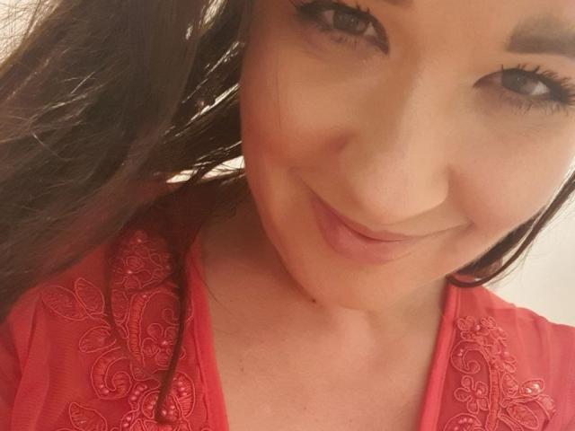 CelestialDevi with a cute smile, wearing a red top with intricate embroidery, has long brown hair and is looking at the camera, showing her face and upper chest.
