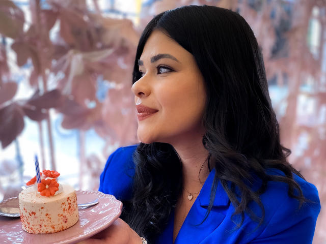 PaulinaValley with long black hair, blue shirt, holding pink plate with white cake, red strawberry, and blue candle, smiling sideways. Her tits are slightly visible under shirt.