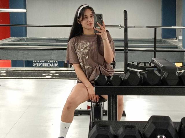 ValeriiNaughty with long black hair, white headband, brown t-shirt, black shorts, white socks, taking a selfie in a gym, sitting on a bench, with dumbbells in front.