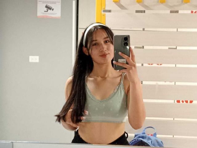 ValeriiNaughty with long black hair, wearing a green sports bra, holding a phone, mirror selfie, showing small tits, flat stomach, and black pants, in a brightly lit room.