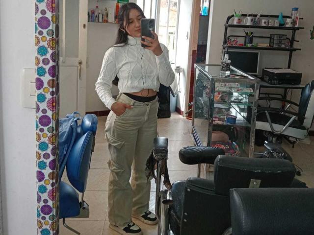 ValeriiNaughty with a cute face, long brown hair, white crop top, tan cargo pants, black belt, black and white shoes, standing in a salon, taking a mirror selfie.