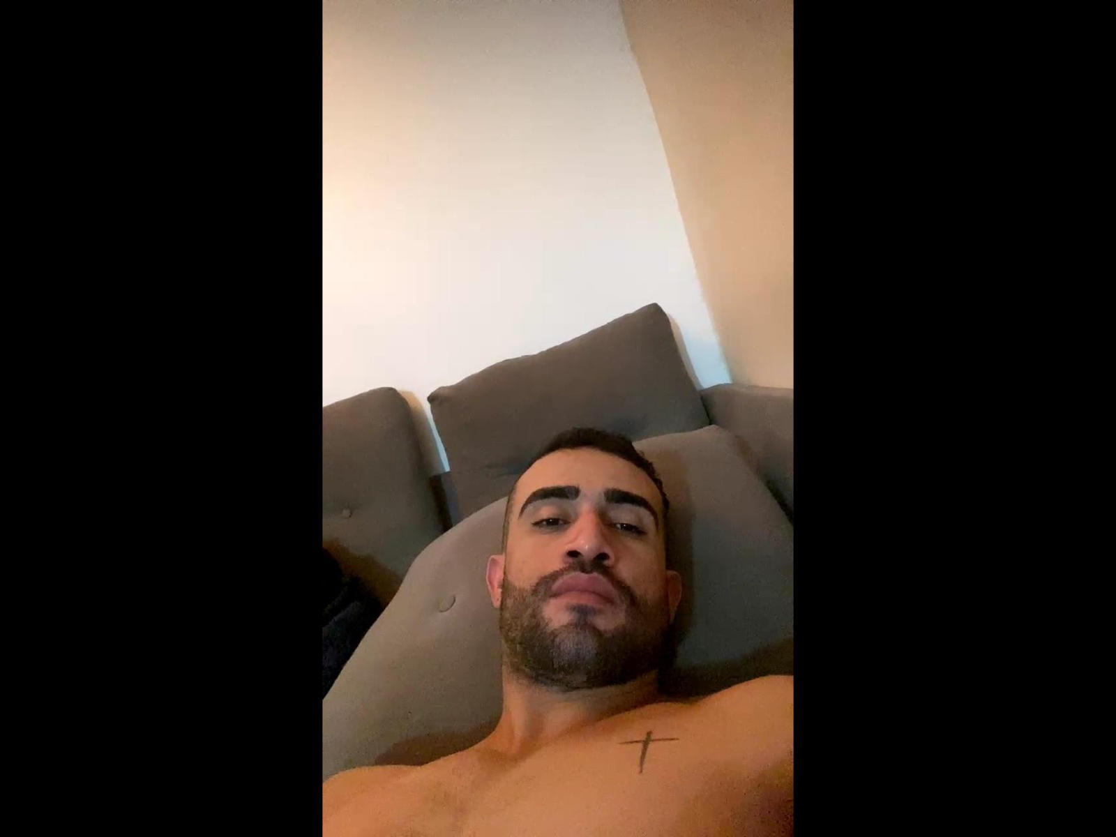 LucasSHot heeft een baard, kort donker haar en een kleine tatoeage van een kruis op zijn rechterborst, ligt zonder shirt met een neutrale uitdrukking en kijkt naar de camera..