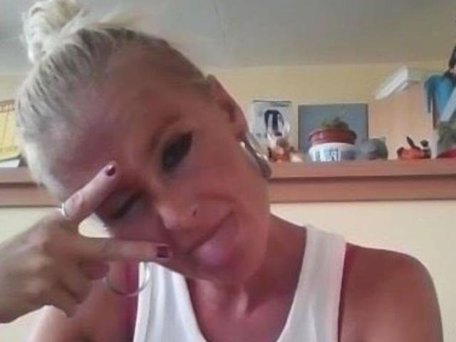 Una ragazza bionda, con una canotta bianca, fa un segno di pace con la mano destra, ha capezzoli rosa, un sorriso appena accennato e smalto rosso sulle unghie, in un selfie leggermente sfocato..
