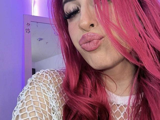 SammyViolet with pink hair, pouty pink lips, white fishnet top, and a nose ring, looking at the camera with her eyes half-open. Her tits are not visible.