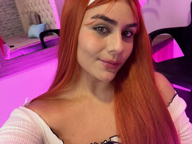 IsabelaCooper with long red hair, white headband, and off-shoulder white top, smiling at the camera, showing her tits slightly, pink lighting in the background.
