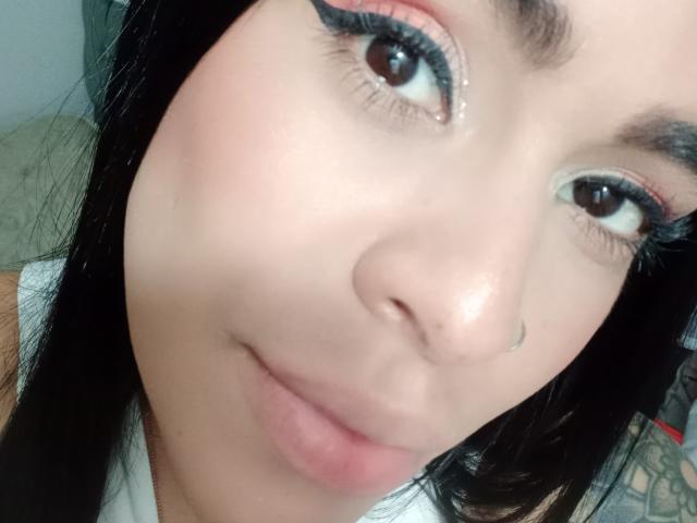 StellaBeckker has light brown skin, black eyeliner, and long black eyelashes. She's smiling with pink lips, a small nose, and a black nose piercing. Her eyes are brown.