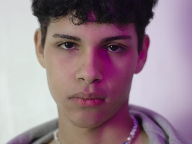 DenisBachh has curly black hair, medium brown skin, and a serious expression. He's wearing a grey hoodie with a white necklace, and there's pink light on his face.