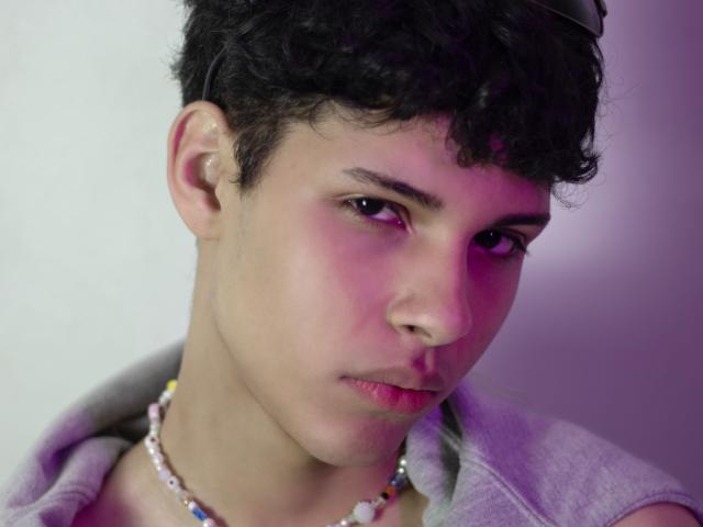 DenisBachh has short curly black hair, a light skin tone, and a serious expression. He's wearing a gray hoodie with a colorful bead necklace. Purple and pink lighting.