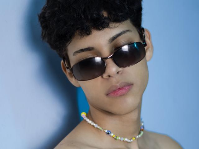 DenisBachh with curly black hair, wearing dark sunglasses, pink lipstick, and a colorful beaded necklace, has a serious expression and a slight head tilt.