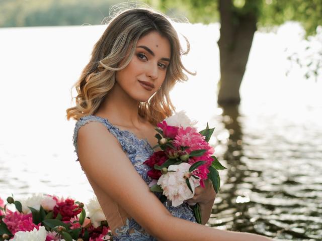 Jasmine, avec ses boucles blondes, portant une robe bleue en dentelle, tenant un bouquet de fleurs rose et blanc, debout dans l'eau, souriant doucement. Ses seins sont discrets, son vagin n'est pas visible..