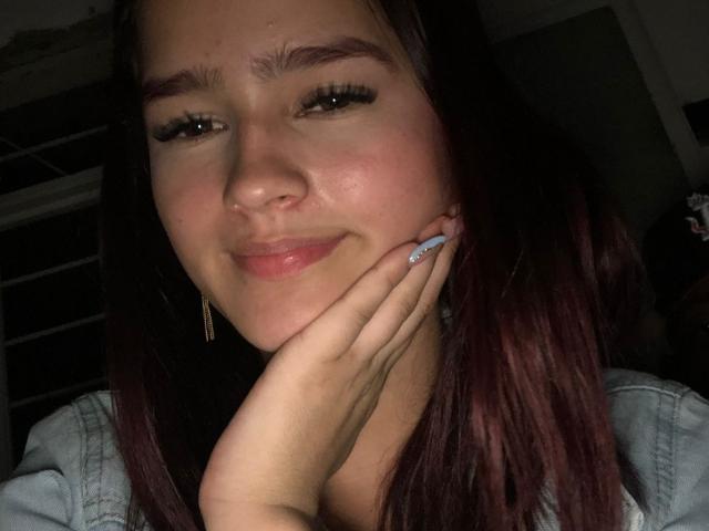 MelodyMansson with dark brown hair, slightly flushed cheeks, and pink glossy lips, smiling softly, touching her face with a hand that has blue and white manicured nails.
