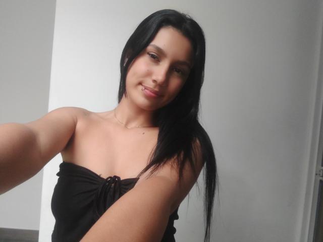 KataleyaMansson, with long black hair, smiling at the camera, wearing a strapless black top, exposing her tits, holding out her arm for a selfie, in a simple room.