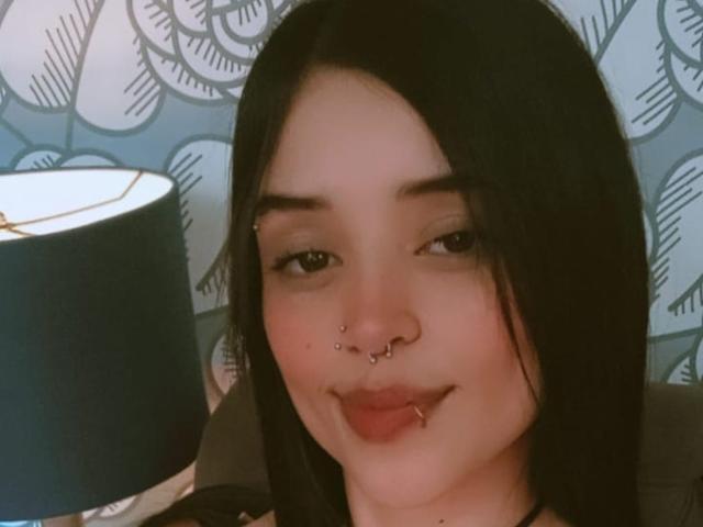Anniuska Taiga, com cabelo preto, piercings no nariz e lábio, e olhos castanhos, está sorrindo levemente, tem um rosto bonito e está olhando para a câmera. Uma luminária está à esquerda..