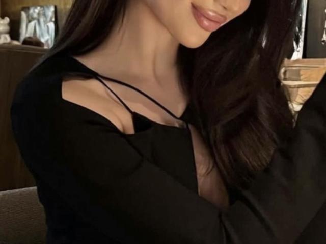 MarinaTender, con lunghi capelli scuri, una maglietta nera che mette in risalto il décolleté e labbra rosa, tiene in mano un oggetto nero e sorride leggermente. Il seno è parzialmente visibile attraverso la maglietta..