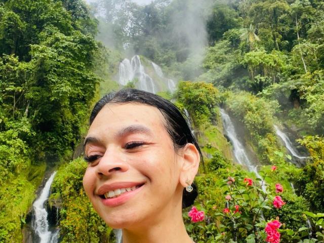 AmberGray69 with dark hair, smiling, showing teeth, and wearing small earrings, in front of a lush green waterfall with mist, and pink flowers in the bottom right.