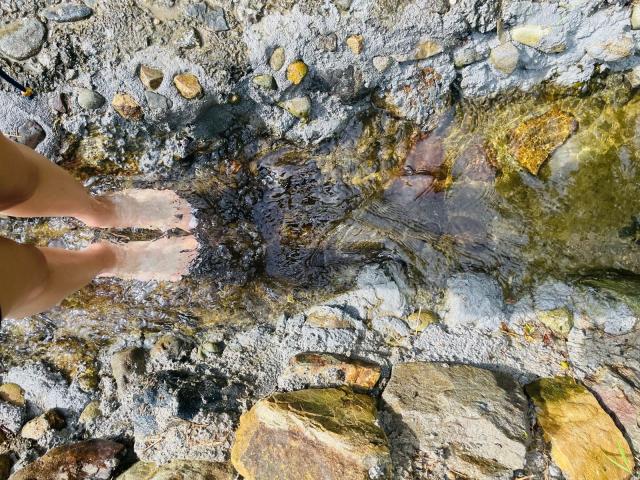 AmberGray69's bare feet in clear water, standing on rocky, pebbled ground with visible stones and small rocks, reflecting sunlight.