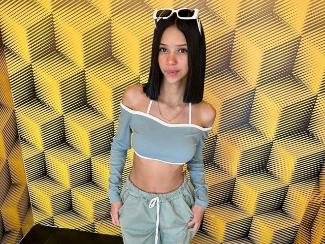 LiaBlom with black straight hair, wearing a blue off-shoulder crop top and grey pants, white sunglasses on head, hands in pockets, showing her small tits and slim ass.