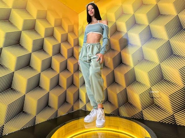 LiaBlom stands in a yellow room wearing a light green crop top and matching high-waisted pants, white sneakers, with hands in pockets, smiling, and showing her flat ass.