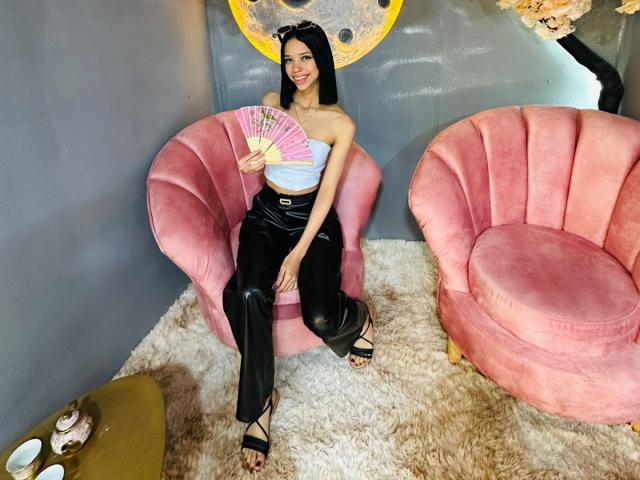 LiaBlom with black hair, smiling, holding pink fan, wearing strapless white top, black leather pants, black sandals, sitting on pink velvet chair, second pink chair to right.